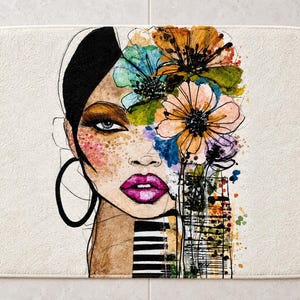May include: A rectangular bath mat featuring a stylized illustration of a face with black hair, large hoop earrings, and bright pink lips. The design incorporates vibrant watercolor flowers in orange, blue, and green, with black ink line details.