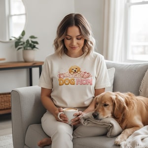 May include: A cream-colored short-sleeve t-shirt featuring a graphic print of a golden puppy resting on a blanket with pink slippers and a mug. The text DOG MOM is printed in large, rounded, multi-colored letters below the puppy illustration.