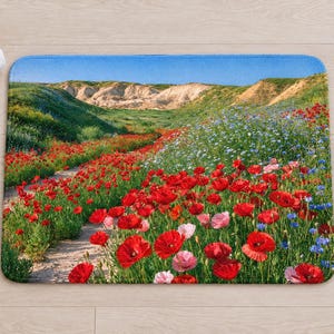 Overhead view of the whole red poppy field bath mat on a light wood floor, with rolled white towels, a lit candle and a small glass vase of wildflowers beside it.