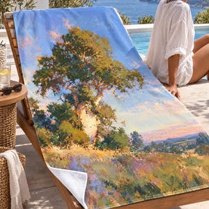 May include: A rectangular beach towel featuring an impressionistic landscape painting of a large tree standing in a field of wildflowers under a blue sky with soft pink clouds. The towel has a white underside and a textured fabric surface.