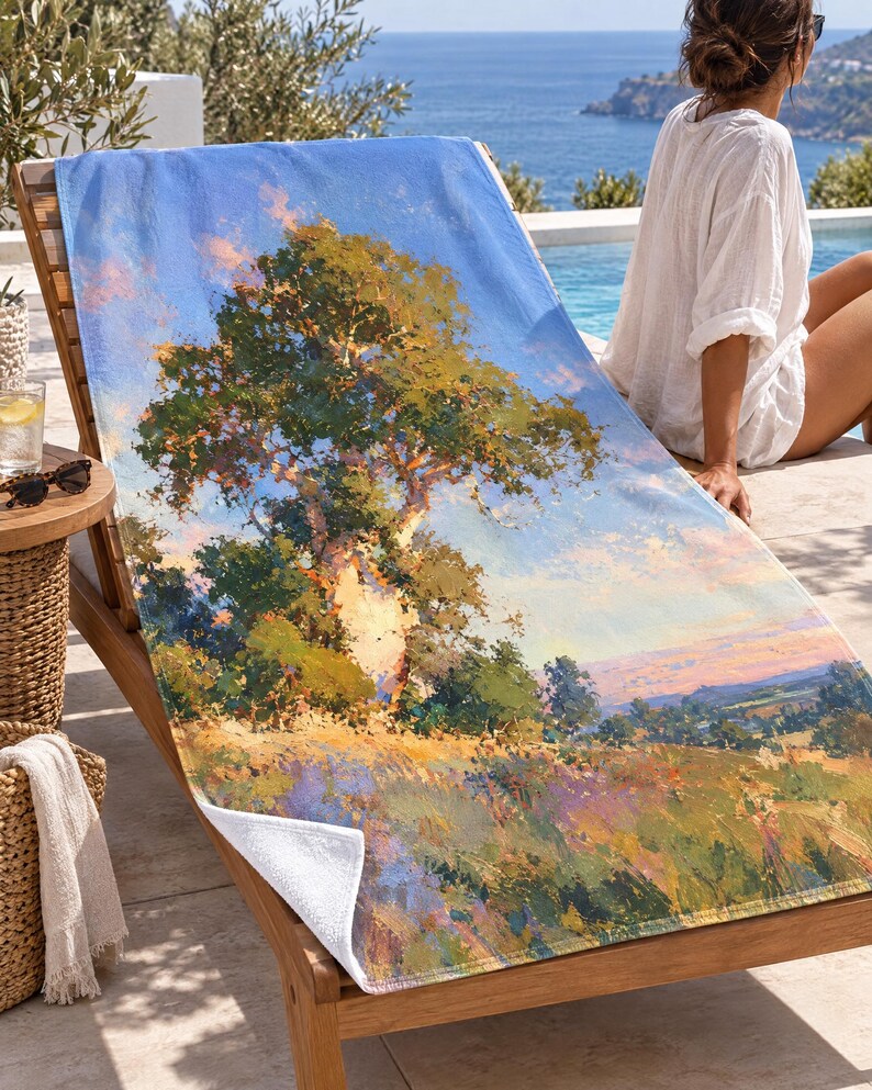May include: A rectangular beach towel featuring an impressionistic landscape painting of a large tree standing in a field of wildflowers under a blue sky with soft pink clouds. The towel has a white underside and a textured fabric surface.