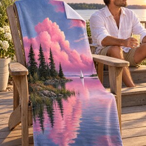 Pine lake beach towel draped on a wooden Adirondack chair on a sunny deck by the water, printed with pink clouds and pine trees reflected in a calm lake with a small sailboat, as a man in a white linen shirt sits nearby.