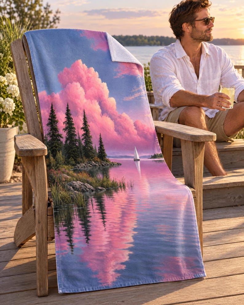 Pine lake beach towel draped on a wooden Adirondack chair on a sunny deck by the water, printed with pink clouds and pine trees reflected in a calm lake with a small sailboat, as a man in a white linen shirt sits nearby.