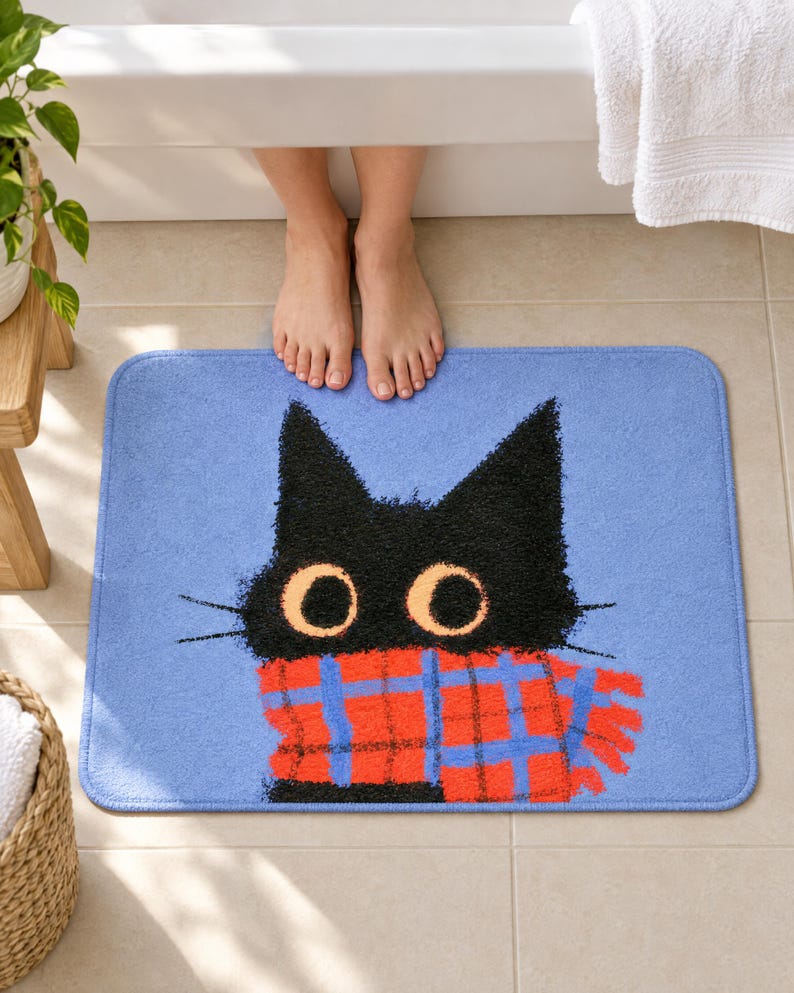Bath mat with a fluffy black cat with big round eyes in a red plaid scarf on periwinkle blue, on a tiled bathroom floor by a tub with bare feet resting on it.
