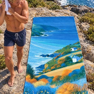 Coastal sea cliff beach towel laid on a rocky shore, printed with blue sea, green cliffs, small white houses and orange wildflowers, as a man in swim shorts dries his hair with a towel beside it.