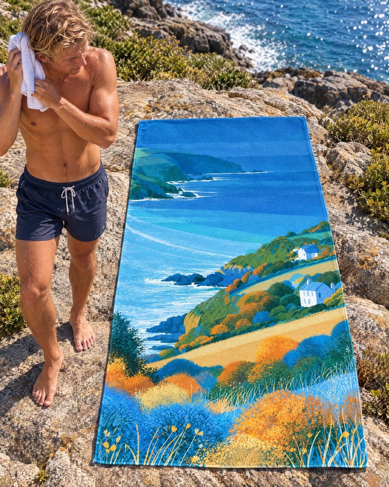 Coastal sea cliff beach towel laid on a rocky shore, printed with blue sea, green cliffs, small white houses and orange wildflowers, as a man in swim shorts dries his hair with a towel beside it.