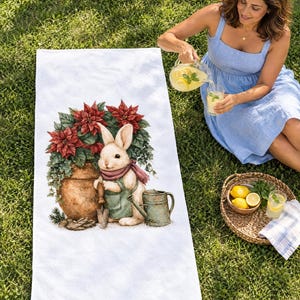 White beach towel printed with a bunny in a green apron beside a potted red poinsettia and watering can, laid flat on sunny grass; a woman sits beside it pouring lemonade.