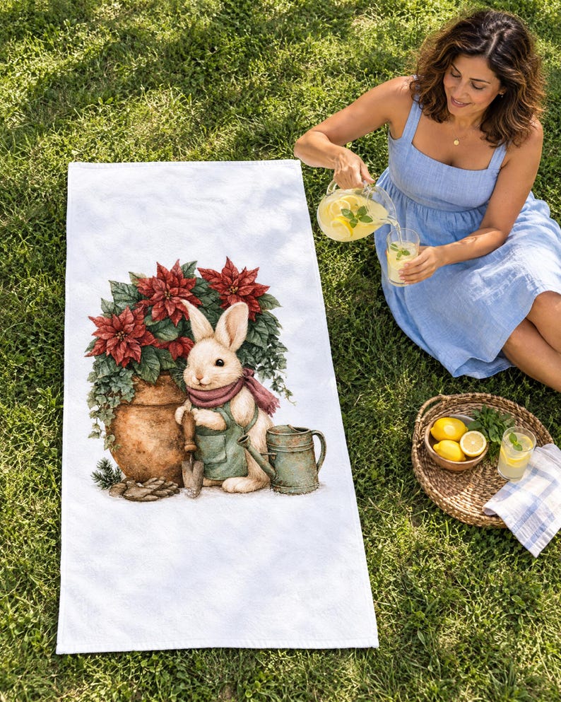 White beach towel printed with a bunny in a green apron beside a potted red poinsettia and watering can, laid flat on sunny grass; a woman sits beside it pouring lemonade.
