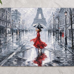 Op de afbeelding: Een rechthoekige badmat met een impressionistisch schilderij van een regenachtige Parijse straat. Een figuur in een felrode jurk houdt een zwarte paraplu vast terwijl deze naar de Eiffeltoren loopt, weerspiegeld in een plas op de grond.