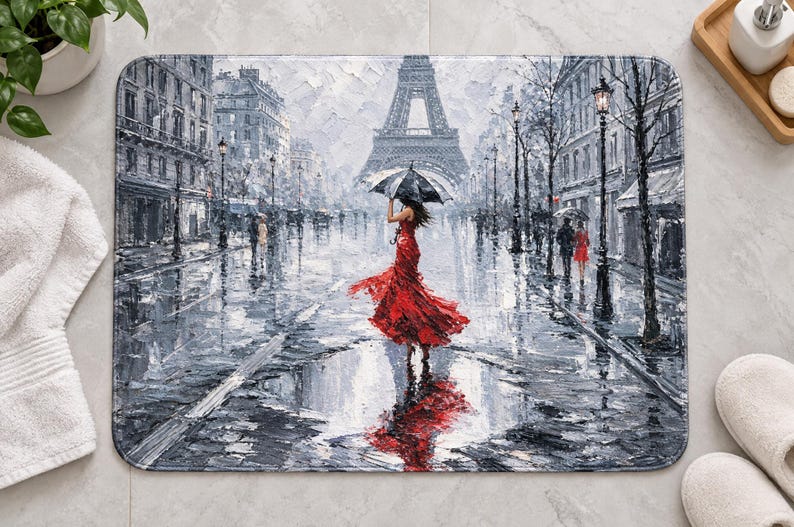 Op de afbeelding: Een rechthoekige badmat met een impressionistisch schilderij van een regenachtige Parijse straat. Een figuur in een felrode jurk houdt een zwarte paraplu vast terwijl deze naar de Eiffeltoren loopt, weerspiegeld in een plas op de grond.