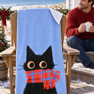 Plaid scarf black cat beach towel draped over a wooden deck chair by the ocean, with a smiling man in a red sweater holding a mug beside it.