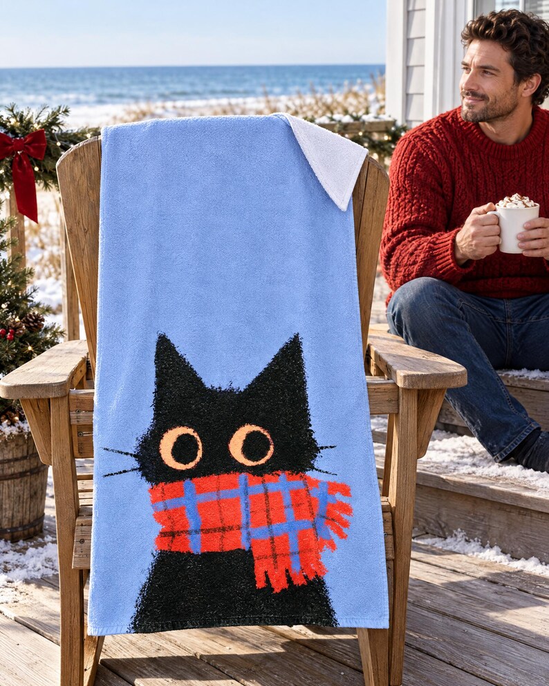 Plaid scarf black cat beach towel draped over a wooden deck chair by the ocean, with a smiling man in a red sweater holding a mug beside it.