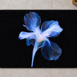 May include: A rectangular bath mat with a solid black background featuring a large, central, ethereal blue flower graphic. The petals display translucent, wispy textures in shades of deep navy, bright cobalt, and soft violet, radiating from a pale centre.