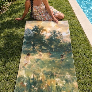 Impressionist garden beach towel laid on green lawn by a pool, printed with soft painted trees, tall grass and small figures in a sunny meadow, as a woman in a floral sundress sits beside it.