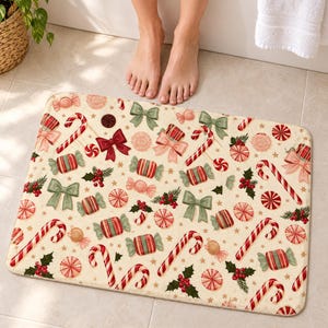 Candy cane Christmas bath mat with candy canes, peppermint swirls, striped candies, bows and holly on cream, on a tiled bathroom floor by a tub with bare feet at the edge.