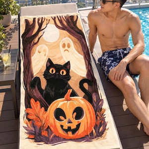 Halloween black cat beach towel on a rooftop pool lounger, printed with a black cat, a carved jack-o'-lantern, ghosts and orange leaves, as a man in sunglasses and swim shorts sits beside it.
