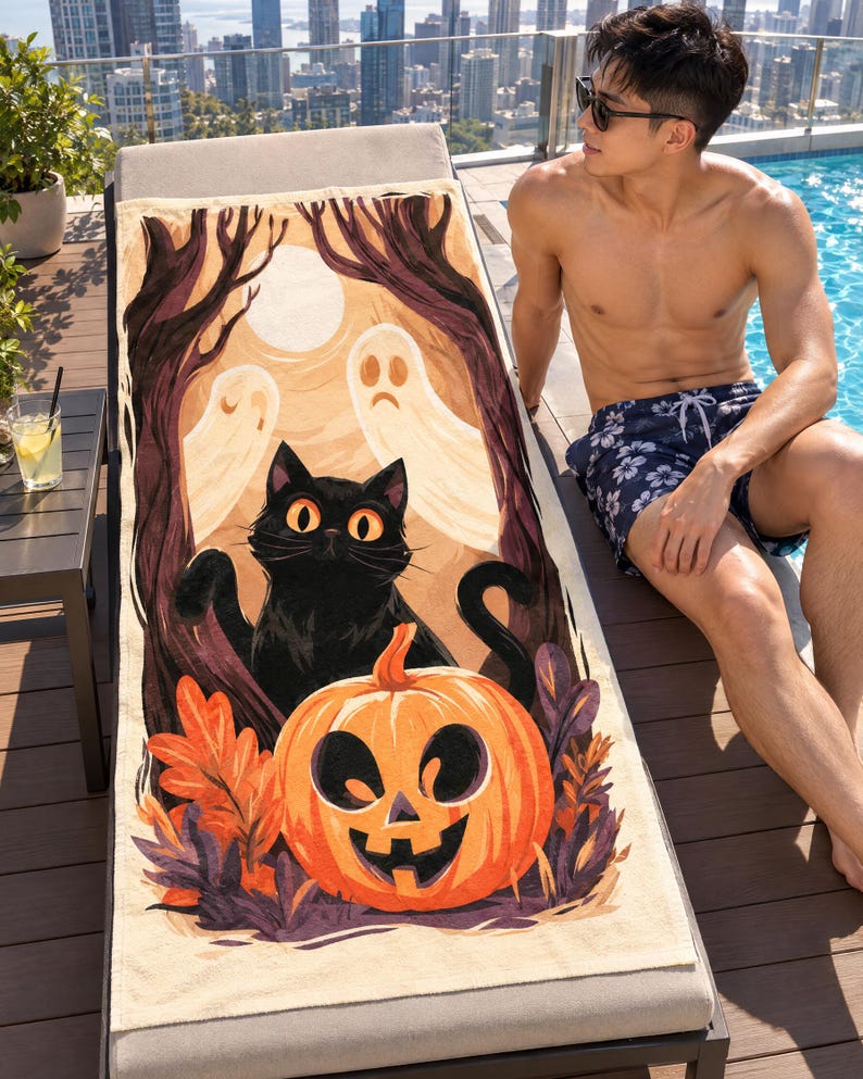 Halloween black cat beach towel on a rooftop pool lounger, printed with a black cat, a carved jack-o'-lantern, ghosts and orange leaves, as a man in sunglasses and swim shorts sits beside it.