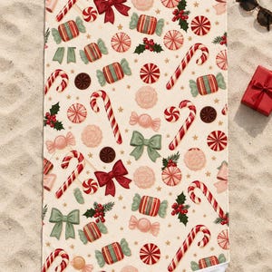 Candy cane Christmas beach towel laid flat on sand with sunglasses and a small red gift box, one corner folded to show the white back.