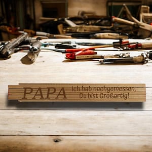 Könnte beinhalten: Ein hölzerner Zollstock mit dem Wort "PAPA" und deutschem Text darauf graviert. Der Zollstock liegt auf einer Holzoberfläche mit verschiedenen Werkzeugen im Hintergrund. Der Text auf dem Zollstock lautet "Ich hab nachgemessen, Du bist Großartig!"