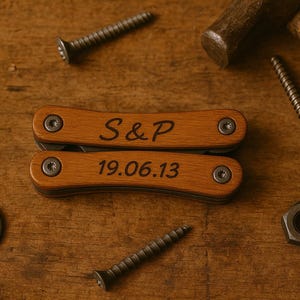 Könnte beinhalten: Ein hölzernes Multitool mit der Gravur "S&P" und "19.06.13" liegt auf einer Holzoberfläche, umgeben von Schrauben, einer Unterlegscheibe, einer Mutter und einem Seil. Das Werkzeug hat eine hellbraune Oberfläche und silberne Beschläge.