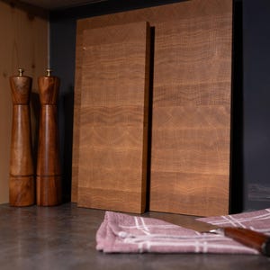 May include: Two wooden cutting boards with a natural wood grain pattern stand upright. Two wooden salt and pepper grinders are to the left. A pink and white checkered cloth and a knife are in the foreground. The items are on a dark countertop.