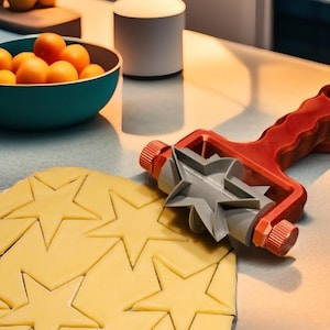 May include: A red and grey star-shaped cookie cutter rolling over dough, creating star shapes. A teal bowl filled with oranges sits nearby. The kitchen scene includes a lamp and a cutting board.