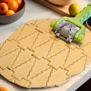 Peut inclure: Un outil de découpe de biscuits vert et gris découpe des formes d'arbres dans une feuille de pâte. La pâte est brun clair et les formes découpées sont visibles. Un bol d'oranges et un citron sont en arrière-plan.