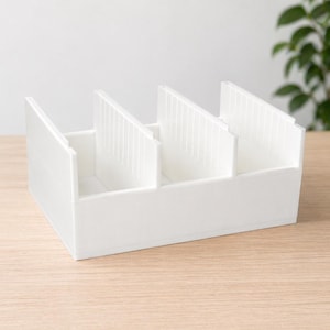 Puede incluir: Un organizador de escritorio rectangular blanco con cuatro compartimentos. El organizador está hecho de un material sólido y opaco y se encuentra sobre una superficie de madera clara. Los divisores tienen una textura acanalada. Una planta verde es visible en el fondo.