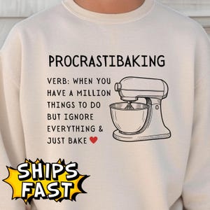 May include: Cream-colored sweatshirt with the text "PROCRASTIBAKING" above a graphic of a stand mixer. The text below reads "VERB: WHEN YOU HAVE A MILLION THINGS TO DO BUT IGNORE EVERYTHING & JUST BAKE".