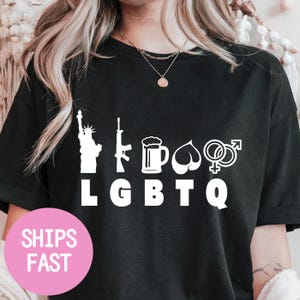 May include: Black t-shirt with white graphic icons including the Statue of Liberty, a rifle, a beer mug, a heart, and gender symbols. The letters "LGBTQ" are printed below the icons. A pink circle with the words "SHIPS FAST" is in the lower left.