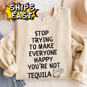 May include: Cream-colored sweatshirt with the text "STOP TRYING TO MAKE EVERYONE HAPPY YOU'RE NOT TEQUILA" in black. A graphic of a cocktail is below the text. The sweatshirt has long sleeves and a crew neck.