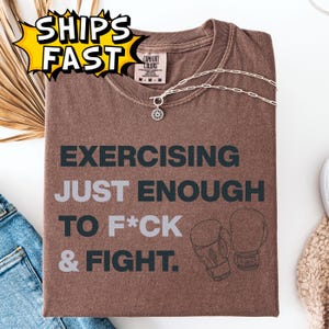 May include: Brown t-shirt with the text "EXERCISING JUST ENOUGH TO F*CK & FIGHT" in dark gray, along with a graphic of boxing gloves. A silver necklace with a flower pendant is on the shirt.