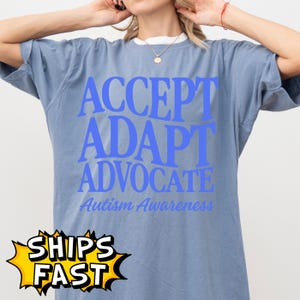 May include: A blue t-shirt with the words "ACCEPT, ADAPT, ADVOCATE" in large blue letters. Below, in a smaller, cursive font, it reads "Autism Awareness". The shirt has a white collar and a "SHIPS FAST" graphic.