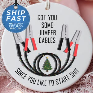 May include: A round, white ceramic Christmas ornament with the text "GOT YOU SOME JUMPER CABLES SINCE YOU LIKE TO START SHIT." The design includes jumper cables and a Christmas tree. A blue circle with "SHIP FAST" is on the left.