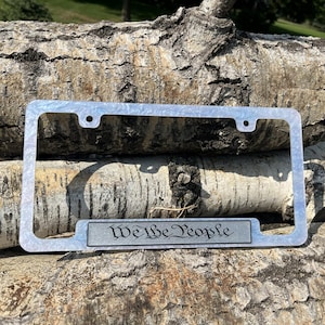 May include: A rectangular license plate frame with a light blue and white marbled finish. The bottom center features a horizontal rectangular plaque with the text "We the People" printed in a black, stylized, traditional script font.