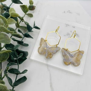 May include: A pair of butterfly earrings. Each earring has a gold-coloured hexagonal frame and a butterfly pendant with a marbled beige, blue, and gold design. The earrings are displayed on a clear, square surface.
