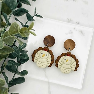 May include: A pair of Thanksgiving-themed earrings. Each earring features a brown turkey design with a cream-colored body, a red wattle, and a wooden stud. The earrings are set against a white background with green foliage.