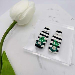 Puede incluir: Pendientes hechos a mano con un diseño de rayas blancas y negras. Cada pendiente tiene forma rectangular y un motivo floral en blanco y verde. Los pendientes se exhiben sobre un bloque de acrílico transparente.