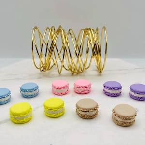 May include: An assortment of colorful macarons in blue, pink, yellow, purple, and tan, with white filling, arranged on a white surface. A gold wire sculpture is behind the macarons. The macarons are a sweet treat.