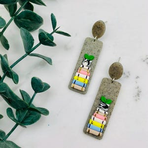 Peut inclure: Une paire de boucles d'oreilles pendantes avec une base rectangulaire gris moucheté, ornée d'une pile de livres colorés rose, jaune, bleu et blanc, surmontée d'une petite feuille verte, suspendue à un clou d'oreille circulaire gris moucheté.