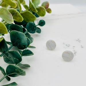 May include: Two small, round, marbled-effect earrings in shades of gray and white are displayed on a white surface. Green and yellow-green leafy sprigs are in the upper left corner. The earrings have a simple, minimalist design.