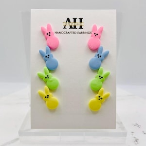 Può includere: Orecchini artigianali a forma di coniglietti nei colori rosa, blu, verde e giallo. Sono esposti su un cartoncino bianco con la scritta "Arts & Hearts HANDCRAFTED EARRINGS".