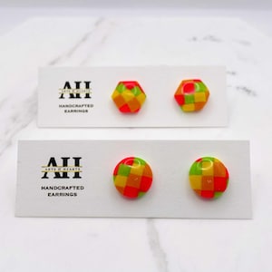 Puede incluir: Dos pares de pendientes hechos a mano sobre tarjetas blancas. Uno es hexagonal, el otro redondo. Ambos presentan un colorido patrón de tablero de ajedrez en rojo, amarillo y verde. Las tarjetas muestran el logo "Arts & Hearts" y el texto "Handcrafted Earrings."