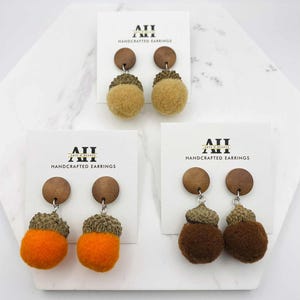 May include: Handcrafted earrings in three styles. Each pair features a wooden top, a textured cap, and a fluffy, round bottom. Colors include tan, orange, and brown. The earring cards display the text "ATT HANDCRAFTED EARRINGS".