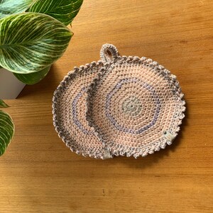 Puede incluir: Un salvamanteles circular de ganchillo hecho a mano en tonos rosa, gris y azul. El salvamanteles tiene un borde festoneado y un lazo para colgar. El diseño presenta círculos concéntricos y está sobre una superficie de madera.