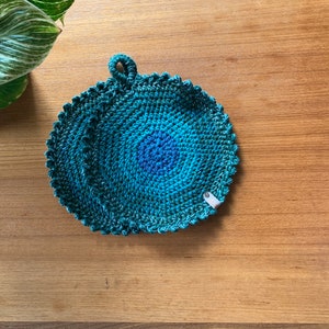 Può includere: Un sottopentola rotondo all'uncinetto con un design a gradiente di blu e verde. Il sottopentola ha un'ansa a forma di anello.