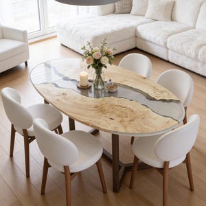 Puede incluir: Mesa de comedor ovalada de madera con un diseño de río de resina transparente, rodeada de seis sillas blancas. Un jarrón con flores y una vela encendida están sobre la mesa. La habitación cuenta con un sofá blanco y una lámpara colgante gris.