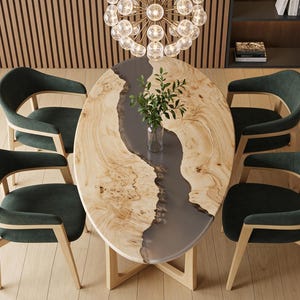 Op de afbeelding: Een ovale eettafel met een oppervlak van natuurlijk licht hout en een centrale inleg van grijze hars, ondersteund door een licht houten onderstel. Vier donkergroene gestoffeerde stoelen met licht houten frames staan rond de tafel.