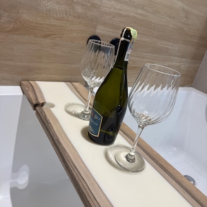 May include: A wooden bathtub tray with a white center holds a green wine bottle and two clear wine glasses. The tray has a light wood border and is set across a white bathtub.