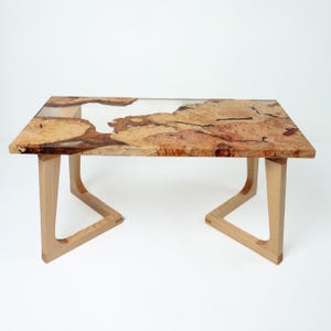 May include: A rectangular wooden coffee table with a clear resin top. The wood has a natural, light brown and reddish-brown burl pattern. The legs are light-colored wood, with a unique angled design. The table is approximately 48 inches long.